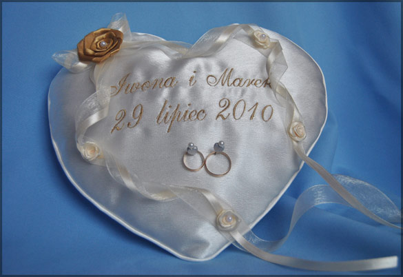 Poduszka w kształcie serca z haftowanym napisem 'Iwona i Marek 29 Lipiec 2010', zdobiona wstążkami i różyczkami, z dwoma złotymi obrączkami umieszczonymi na środku.