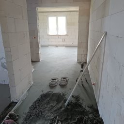 Piotrowski Okna i Drzwi - Etap prac posadzkarskich: świeżo wylana wylewka podłogowa w pomieszczeniu z białych bloczków, z widoczną kupą mokrej mieszanki, łatą i laserem budowlanym. Na podłodze klapki robocze, w tle okno.