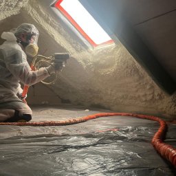 Piotrowski Okna i Drzwi - Pracownik w kombinezonie i masce natryskuje piankę PUR na skośne ściany poddasza, obok świetlika dachowego. Podłoga zabezpieczona ciemną folią, widoczny pomarańczowy wąż.
