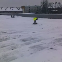 Pracownik w żółtej kamizelce na płaskim dachu pokrytym śniegiem, w tle budynki miejskie w pochmurny dzień.