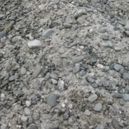 Góra szarego kruszywa o zróżnicowanej frakcji, od drobnego piasku po zaokrąglone kamienie o średnicy kilku centymetrów, tworzące niejednolitą teksturę.