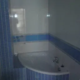 Narożna wanna obudowana niebieską mozaiką, białe płytki na ścianach z niebieskim paskiem dekoracyjnym, widoczna armatura i fragment toalety.