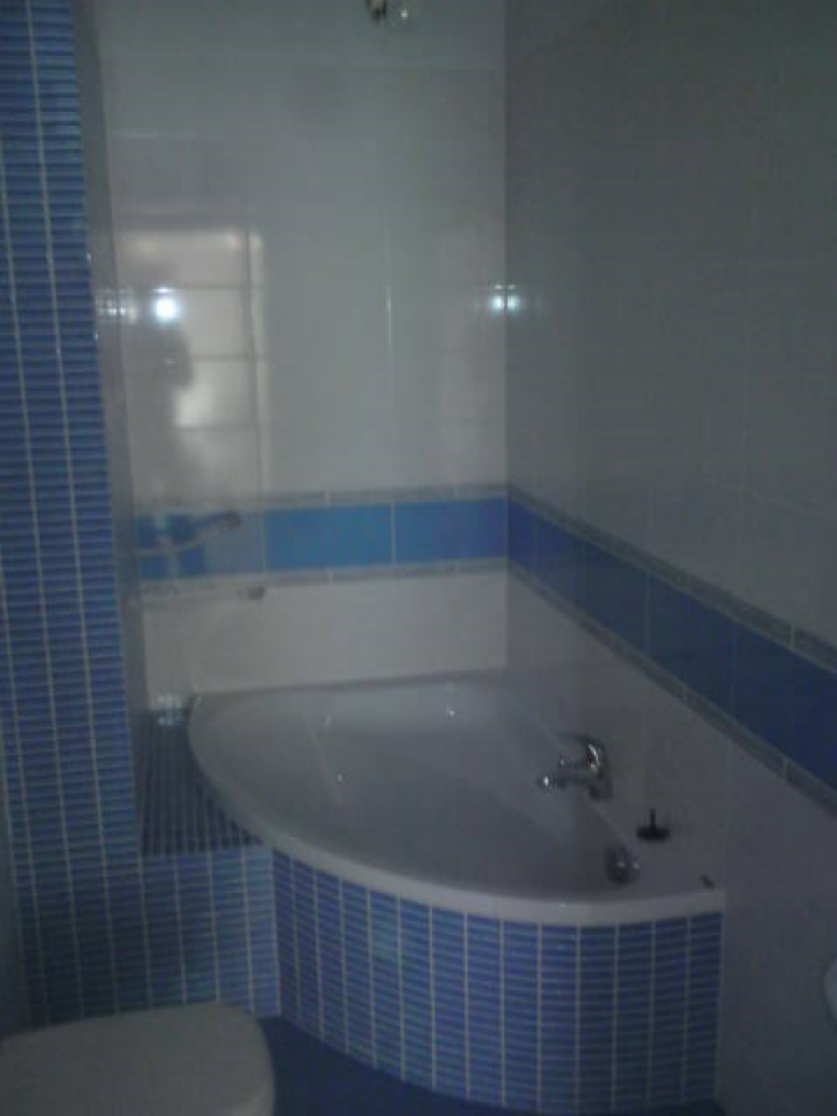 Narożna wanna obudowana niebieską mozaiką, białe płytki na ścianach z niebieskim paskiem dekoracyjnym, widoczna armatura i fragment toalety.