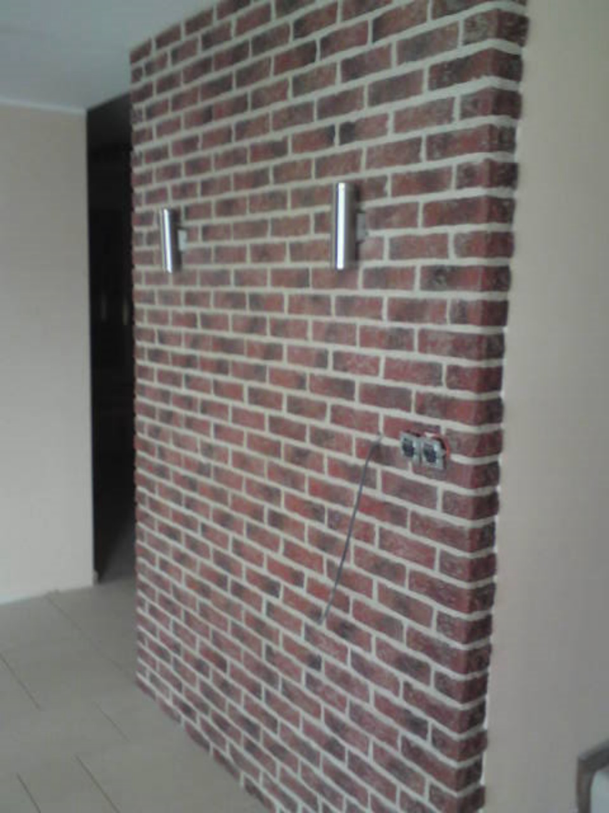 Ściana wykończona czerwoną cegłą klinkierową z białymi fugami, oświetlona dwoma srebrnymi kinkietami, z widocznym gniazdkiem elektrycznym z wystającym kablem.