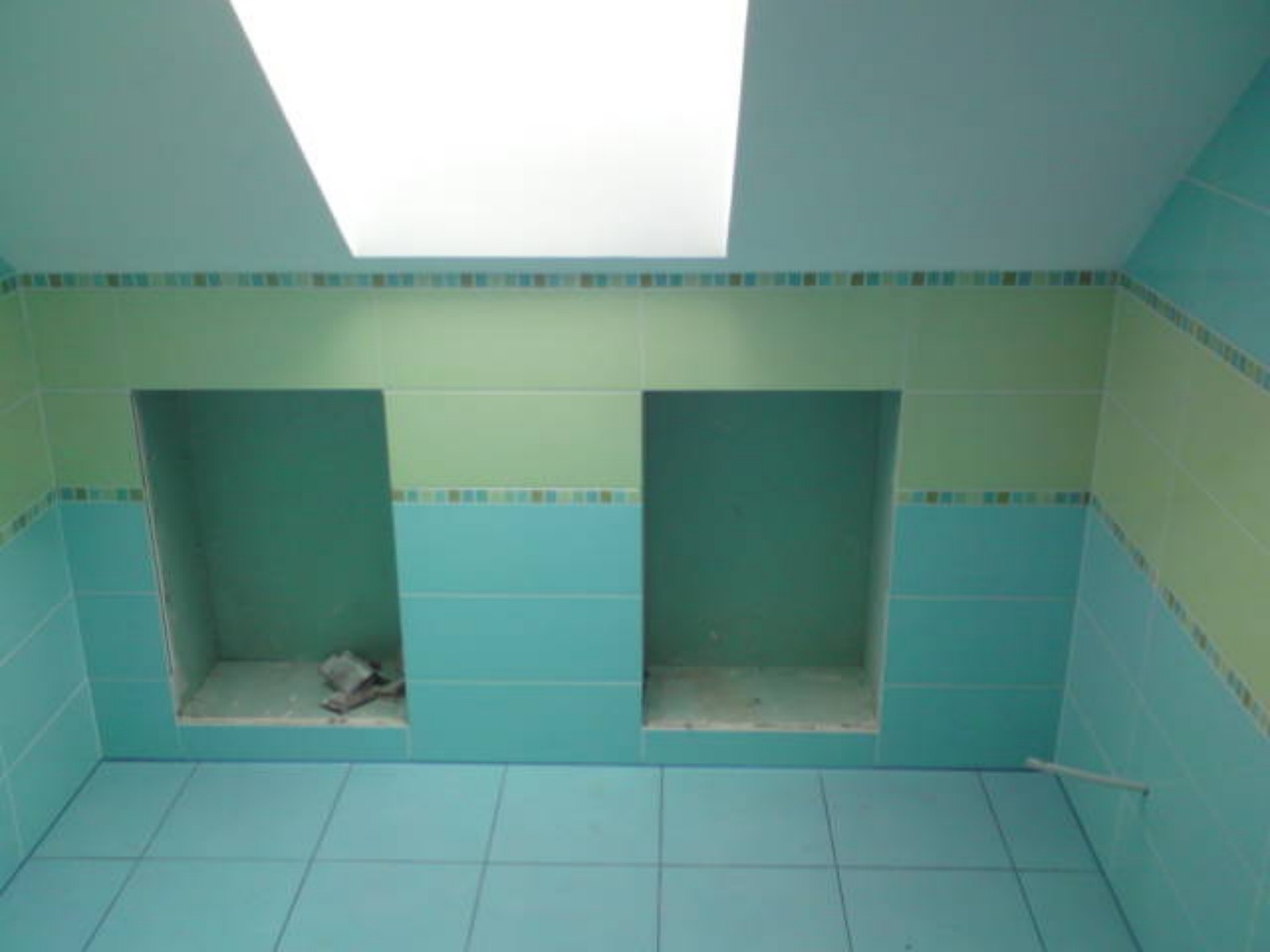 Łazienka w trakcie wykańczania, z turkusowymi i zielonymi płytkami, widoczne dwie wnęki w ścianie, sufit z oknem dachowym, na podłodze płytki w kolorze turkusowym.