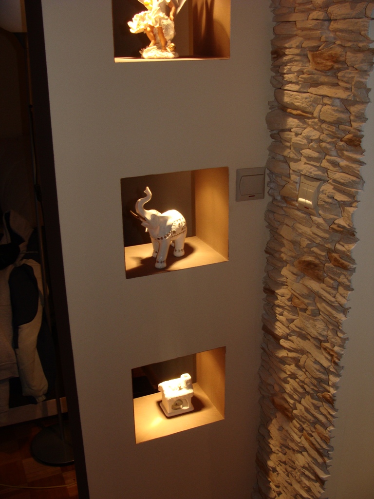 Ściana z trzema wnękami podświetlonymi ciepłym światłem, w których stoją ozdobne figurki i kolumna wyłożona kamieniem dekoracyjnym.