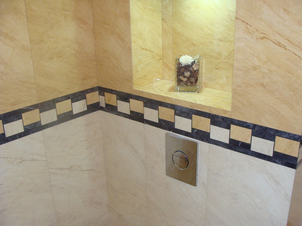 Wykończona ściana w łazience z beżowych płytek marmurowych, ozdobiona dekoracyjnym pasem z małych, kwadratowych płytek w odcieniach beżu, bieli i szarości, z widocznym przyciskiem spłuczki toalety...