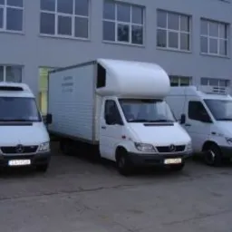 Trzy białe busy dostawcze zaparkowane przed szarym budynkiem. Środkowy bus z zabudową kontenerową. Tablice rejestracyjne widoczne na każdym pojeździe. Ujęcie z poziomu gruntu, lekko z lewej strony.