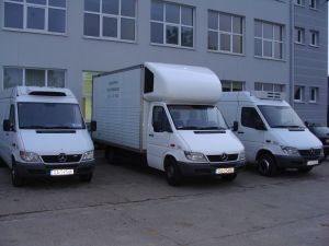 Trzy białe busy dostawcze zaparkowane przed szarym budynkiem. Środkowy bus z zabudową kontenerową. Tablice rejestracyjne widoczne na każdym pojeździe. Ujęcie z poziomu gruntu, lekko z lewej strony.