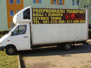Biały samochód dostawczy z napisem 'Przeprowadzki Transport Kraj i Europa Szybko Tanio' oraz numerami telefonów, zaparkowany na osiedlu mieszkaniowym w słoneczny dzień.