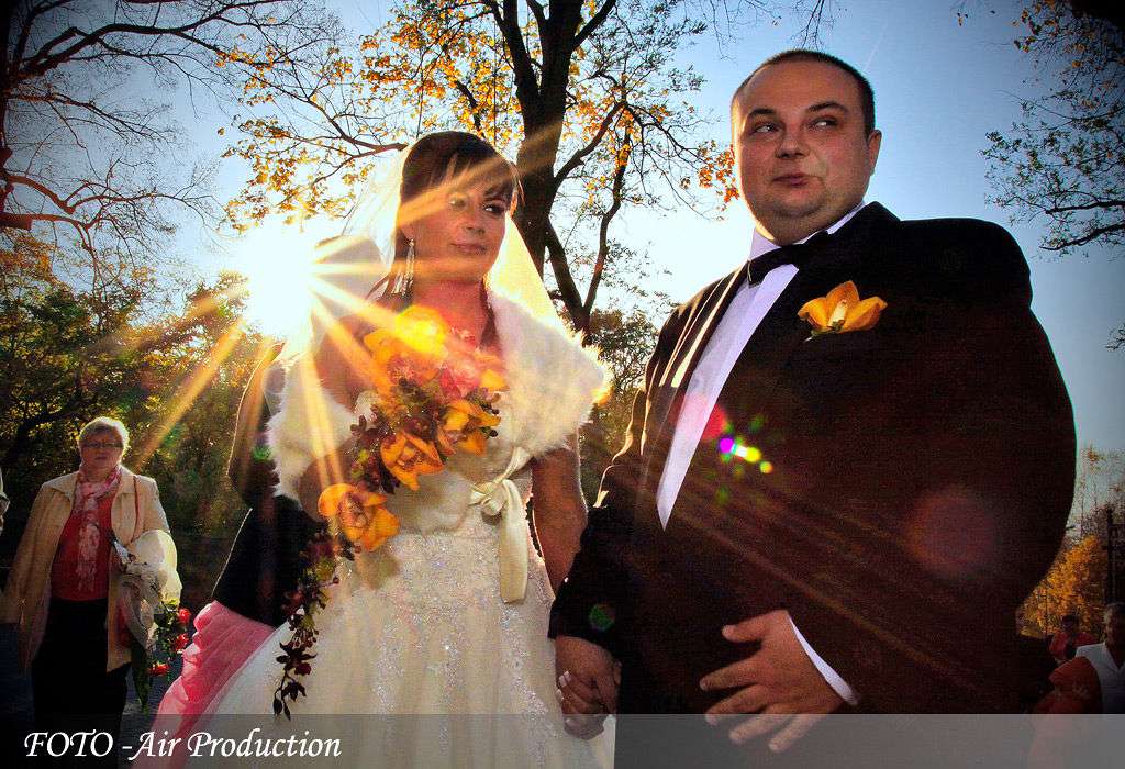 Para młoda trzymająca się za ręce, w tle jesienne drzewa i słońce, widoczna starsza kobieta z kwiatami.