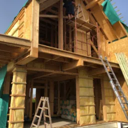 Wznoszenie konstrukcji domu z drewnianych belek, widoczne ocieplenie ścian wełną mineralną, na górze pracownik na rusztowaniu, zabezpieczenie dachu zieloną folią.