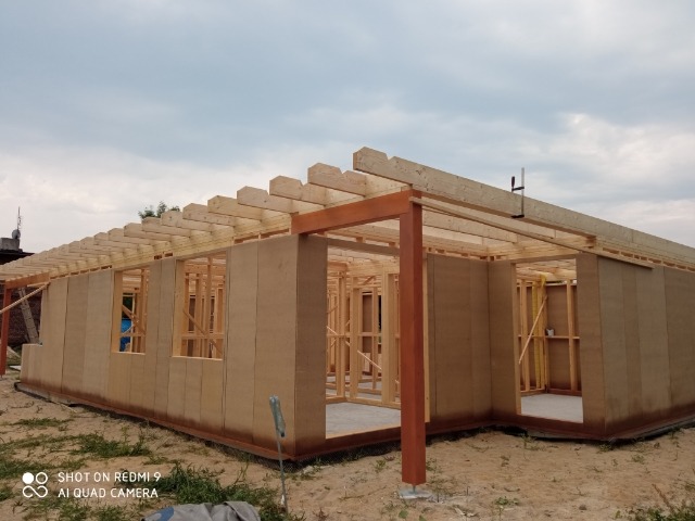 Wznoszenie konstrukcji parterowego domu z drewnianym szkieletem, widoczne ściany z płyt i elementy więźby dachowej.