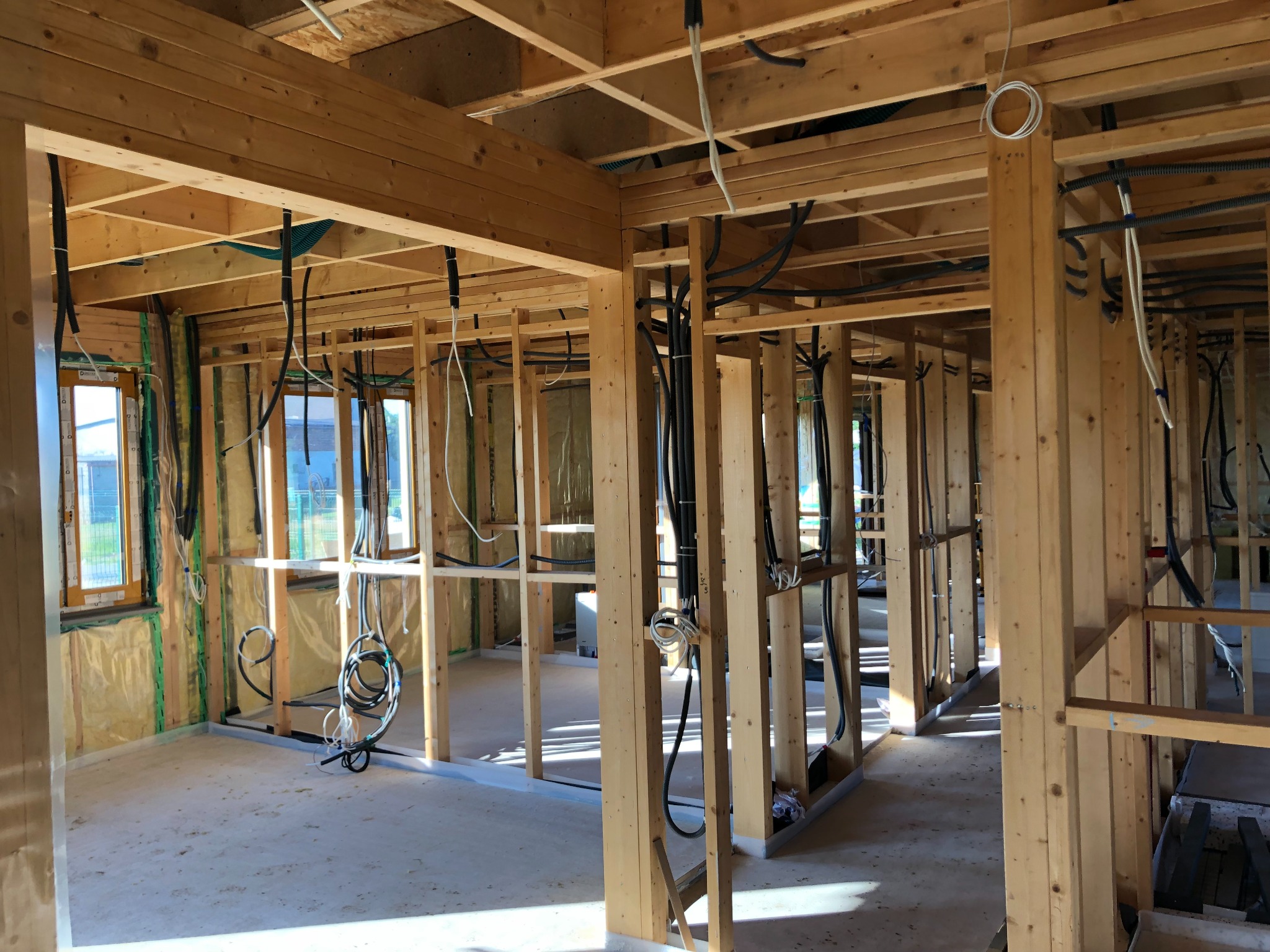 Wnętrze w trakcie budowy, widoczne drewniane elementy konstrukcyjne, instalacje elektryczne oraz okna z widokiem na zewnątrz.