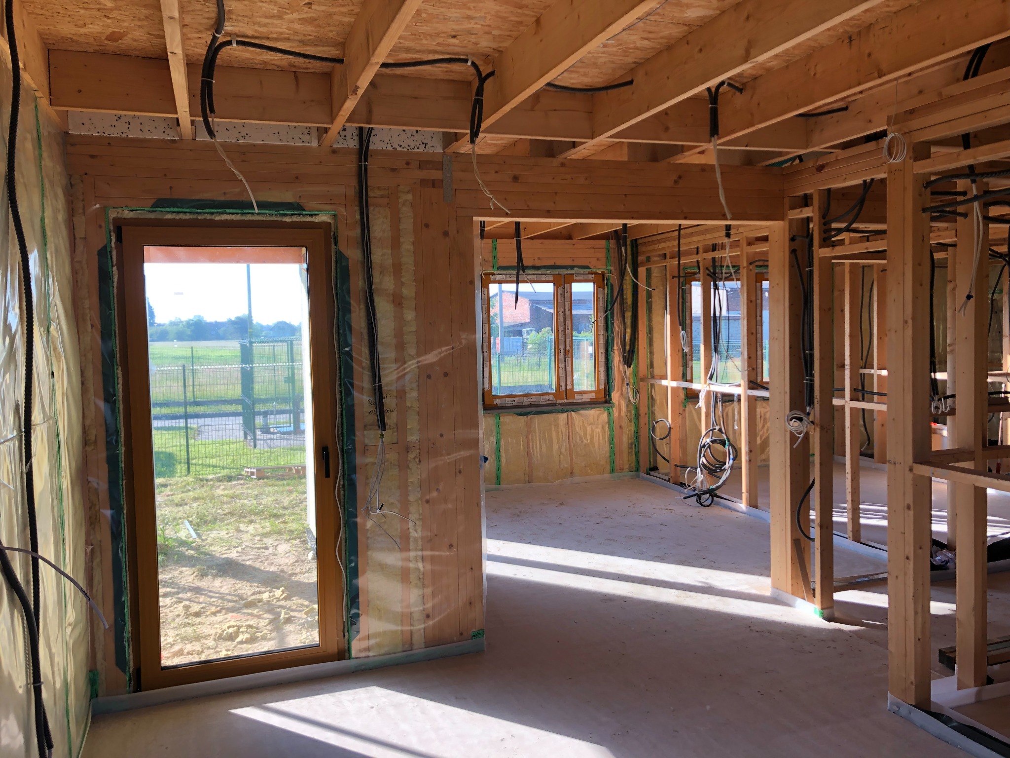 Wnętrze w stanie surowym otwartym z widoczną konstrukcją drewnianą, instalacją elektryczną, oknami i drzwiami, folią ochronną na ścianach i wylewką podłogową.