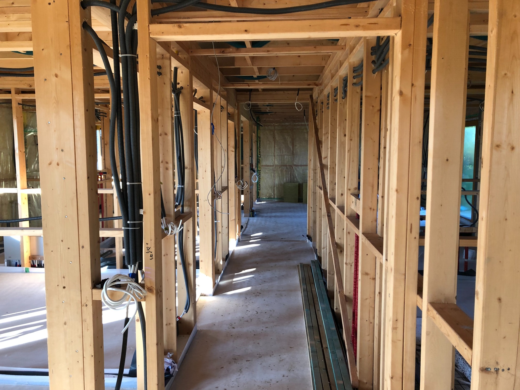 Wnętrze w trakcie budowy: drewniany szkielet domu z widocznymi instalacjami elektrycznymi i sanitarnymi, rzut oka na korytarz z elementami izolacji w tle.