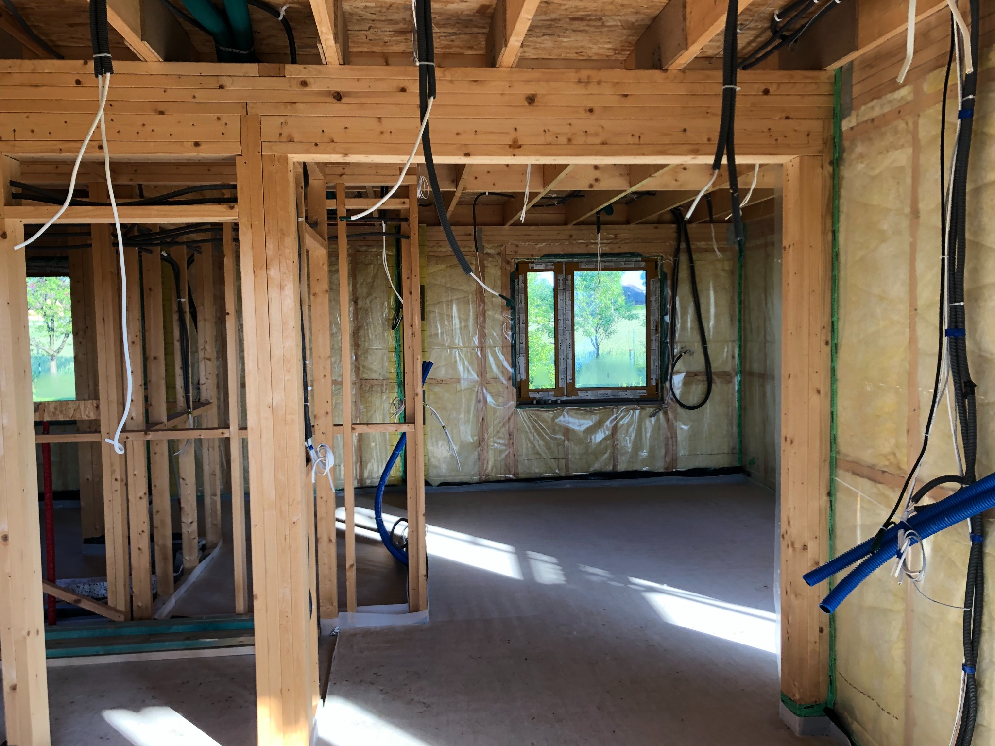 Wnętrze w trakcie budowy: drewniany szkielet domu z widoczną izolacją, instalacje elektryczne i hydrauliczne, okno z widokiem na zieleń.