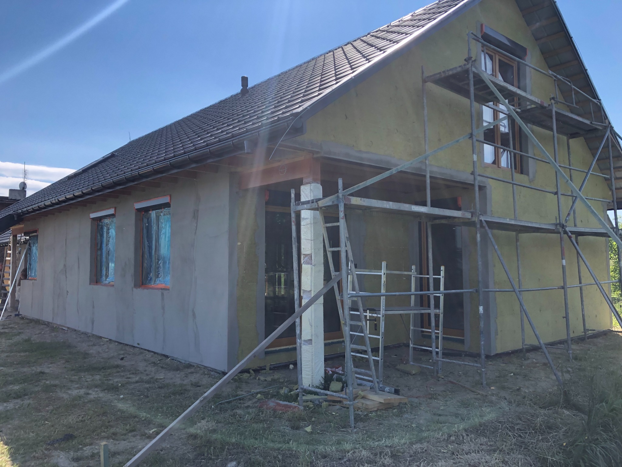 Budowa domu szkieletowego z ociepleniem zewnętrznym i częściowo otynkowanymi ścianami, rusztowania przy elewacji, okna zabezpieczone folią.