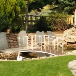 Ogród z małym stawem, białym mostkiem i kaskadą wodną z kamienia, otoczony zielenią i ozdobnym ogrodzeniem.