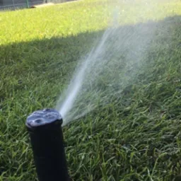 Automatyczny zraszacz nawadnia trawnik w Prószkowie, w tle nowoczesny budynek mieszkalny i ogrodzenie.