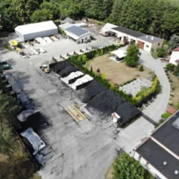 Widok z lotu ptaka na skład opału z hałdami węgla, zadaszonym miejscem parkingowym i budynkami magazynowymi, w tle las