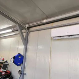 Biały klimatyzator Gree zamontowany w hali produkcyjnej lub magazynie, widoczna instalacja elektryczna i niebieski zwijak do węża.