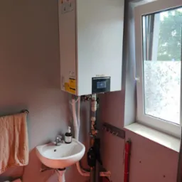 Biała pompa ciepła marki Gree zamontowana na szarej ścianie nad umywalką w małej łazience z oknem z ozdobną folią.