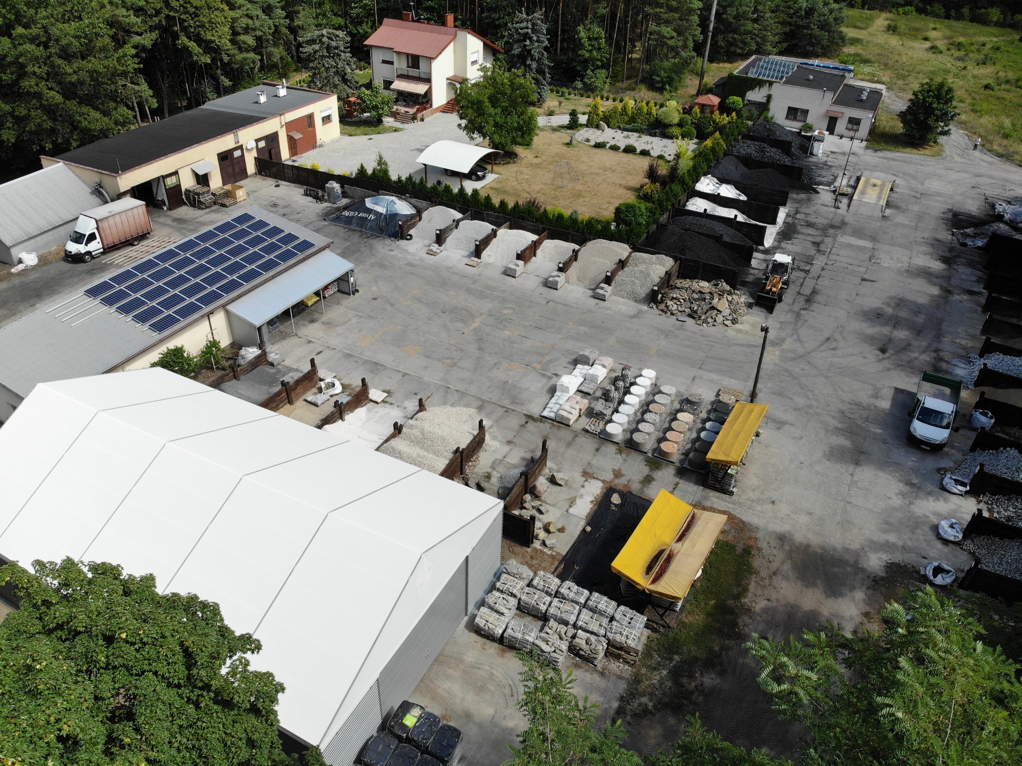 Widok z góry na skład materiałów budowlanych z różnymi rodzajami kruszyw, węgla i kamieni dekoracyjnych, składowanych w oddzielnych boksach i na paletach, z budynkami w tle i panelami słonecznymi...