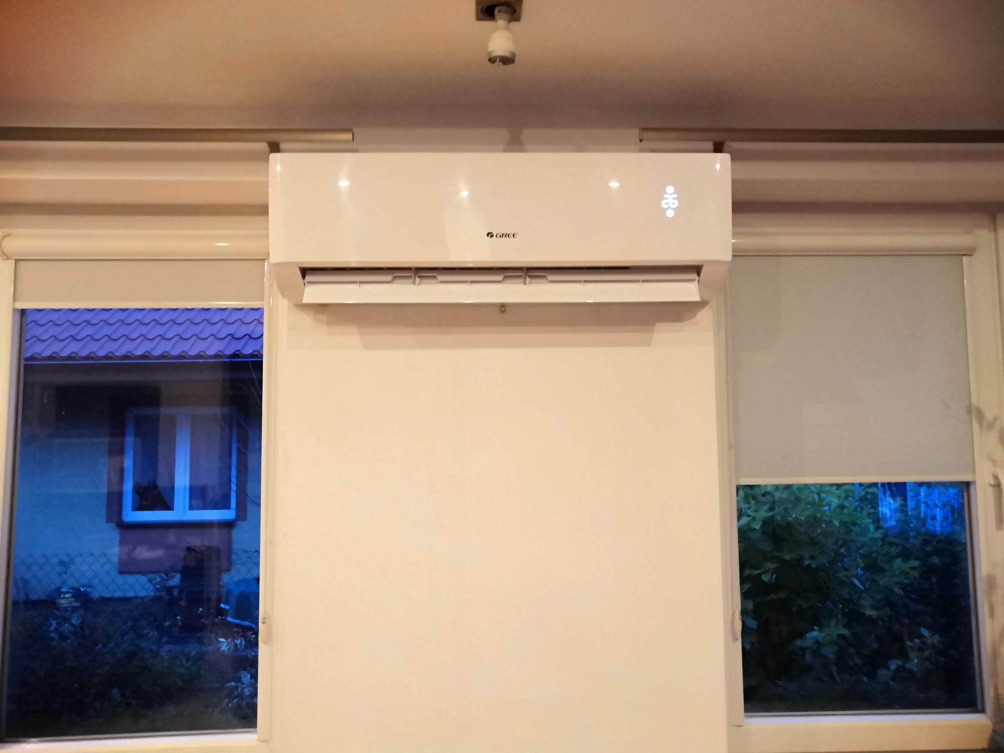 Biały klimatyzator marki Gree zamontowany na ścianie w pokoju, widok z przodu, okna z roletami po bokach, widoczny wyświetlacz temperatury.