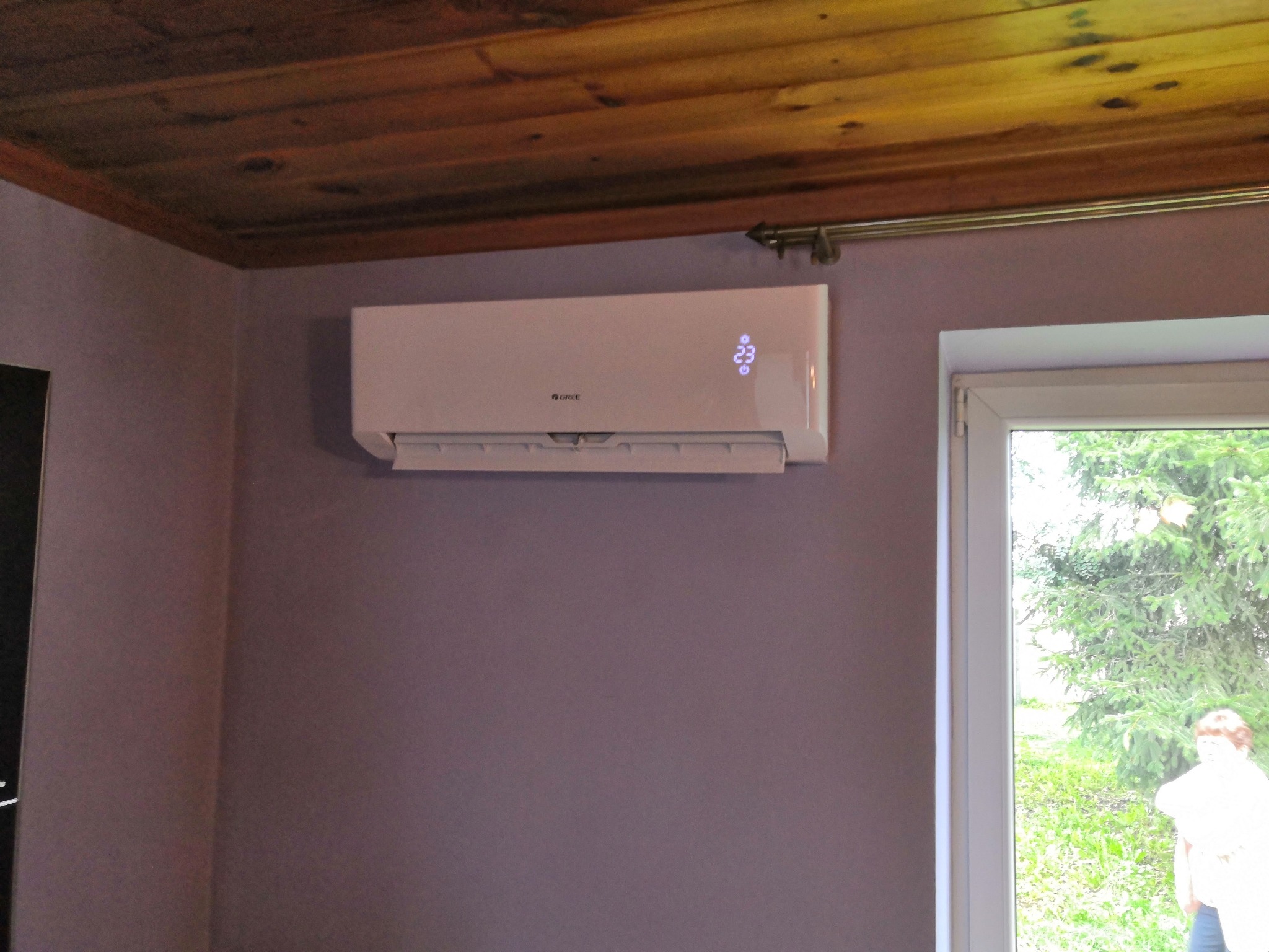 Biały klimatyzator z wyświetlaczem temperatury (23 stopnie Celsjusza) zamontowany na fioletowej ścianie w pokoju z drewnianym sufitem, widok przez okno na zieleń na zewnątrz.