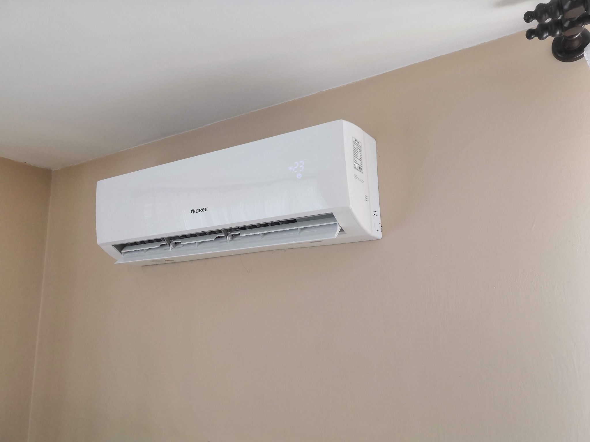 Biała klimatyzacja marki Gree zamontowana na beżowej ścianie w pomieszczeniu, wyświetlająca temperaturę 23 stopnie Celsjusza.