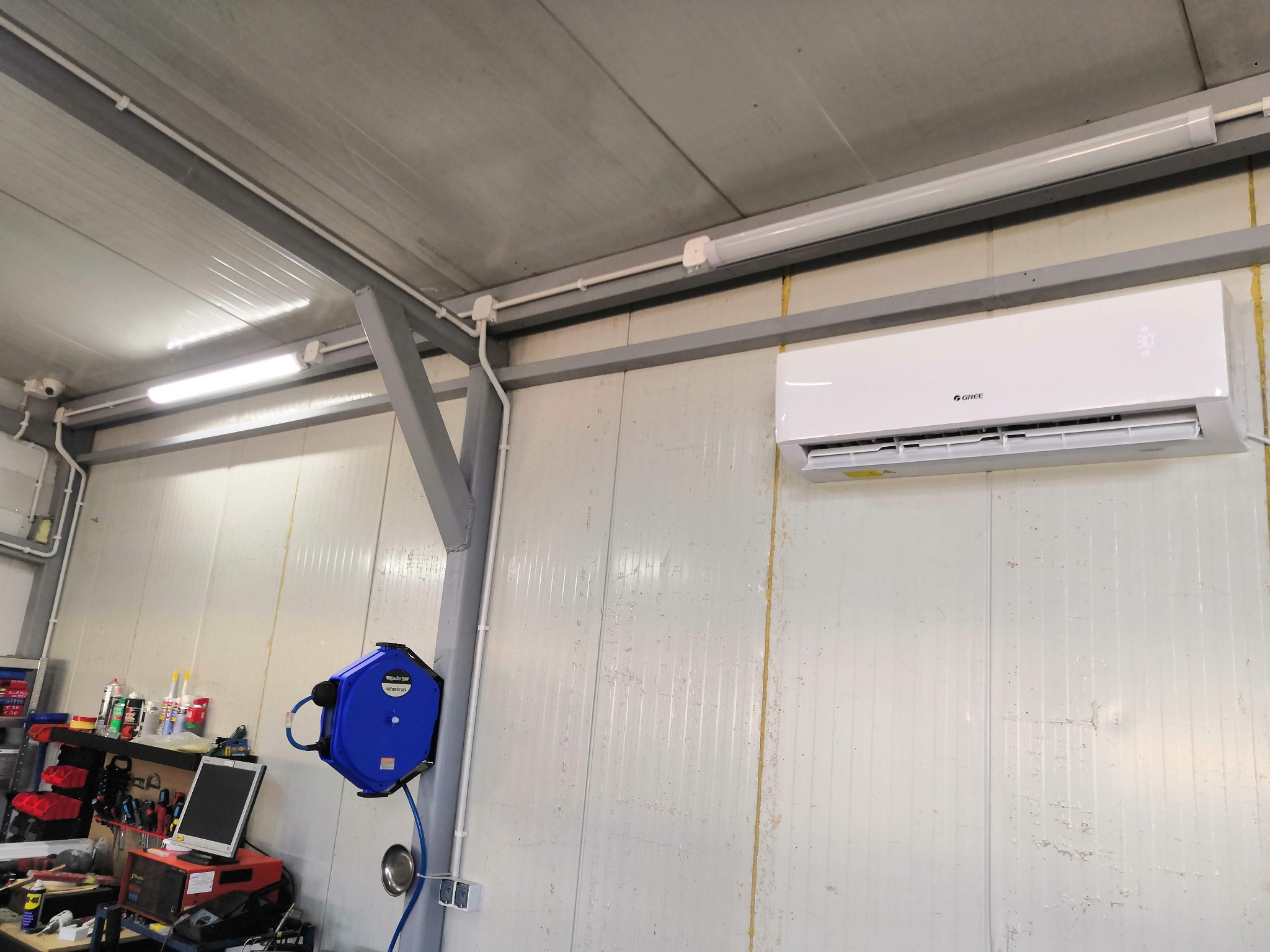 Biały klimatyzator Gree zamontowany w hali produkcyjnej lub magazynie, widoczna instalacja elektryczna i niebieski zwijak do węża.