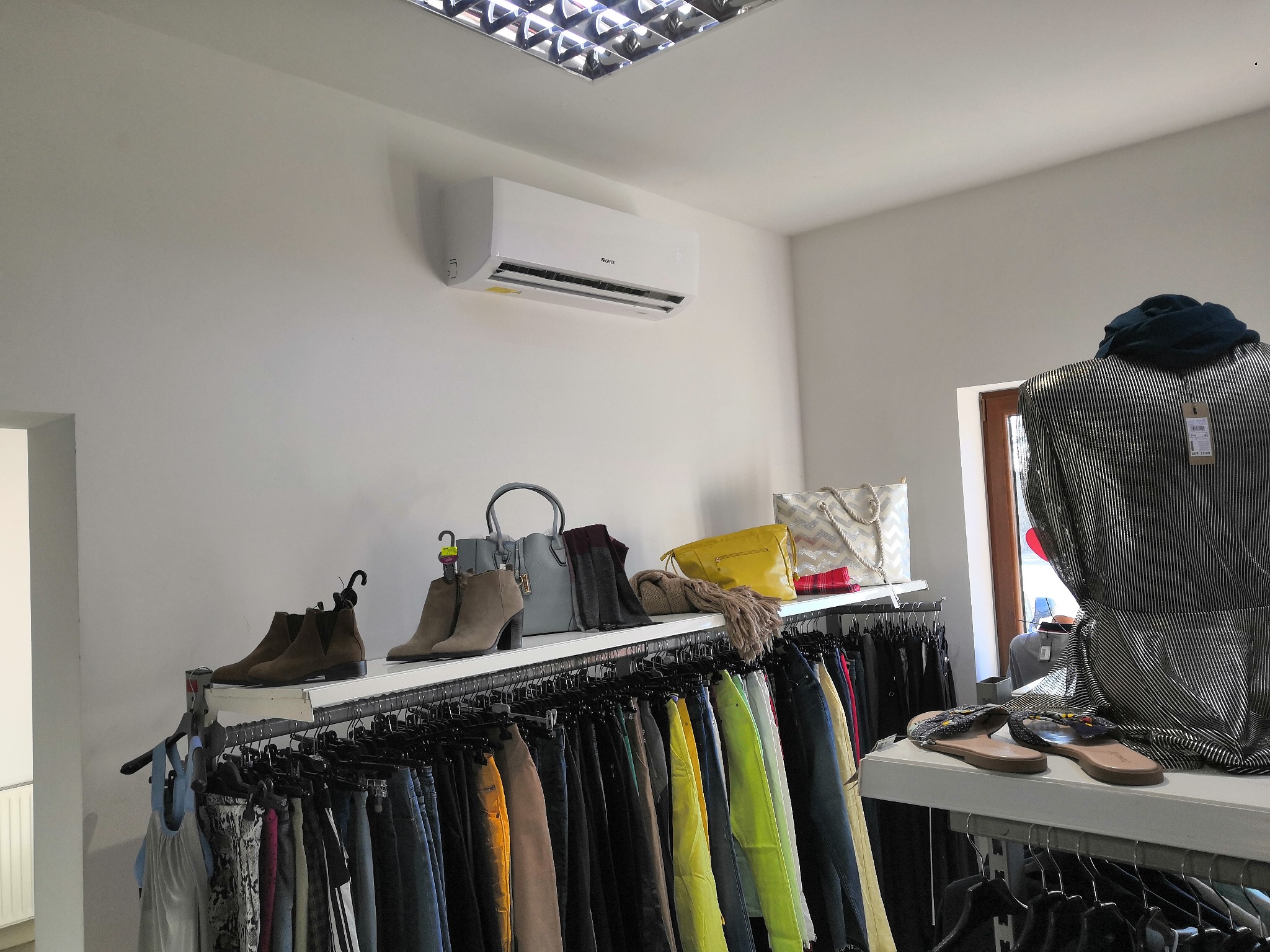 Biały klimatyzator marki Gree zamontowany na ścianie w butiku odzieżowym, widoczne ubrania na wieszakach i torebki na półce, oświetlenie sufitowe rastrowe.
