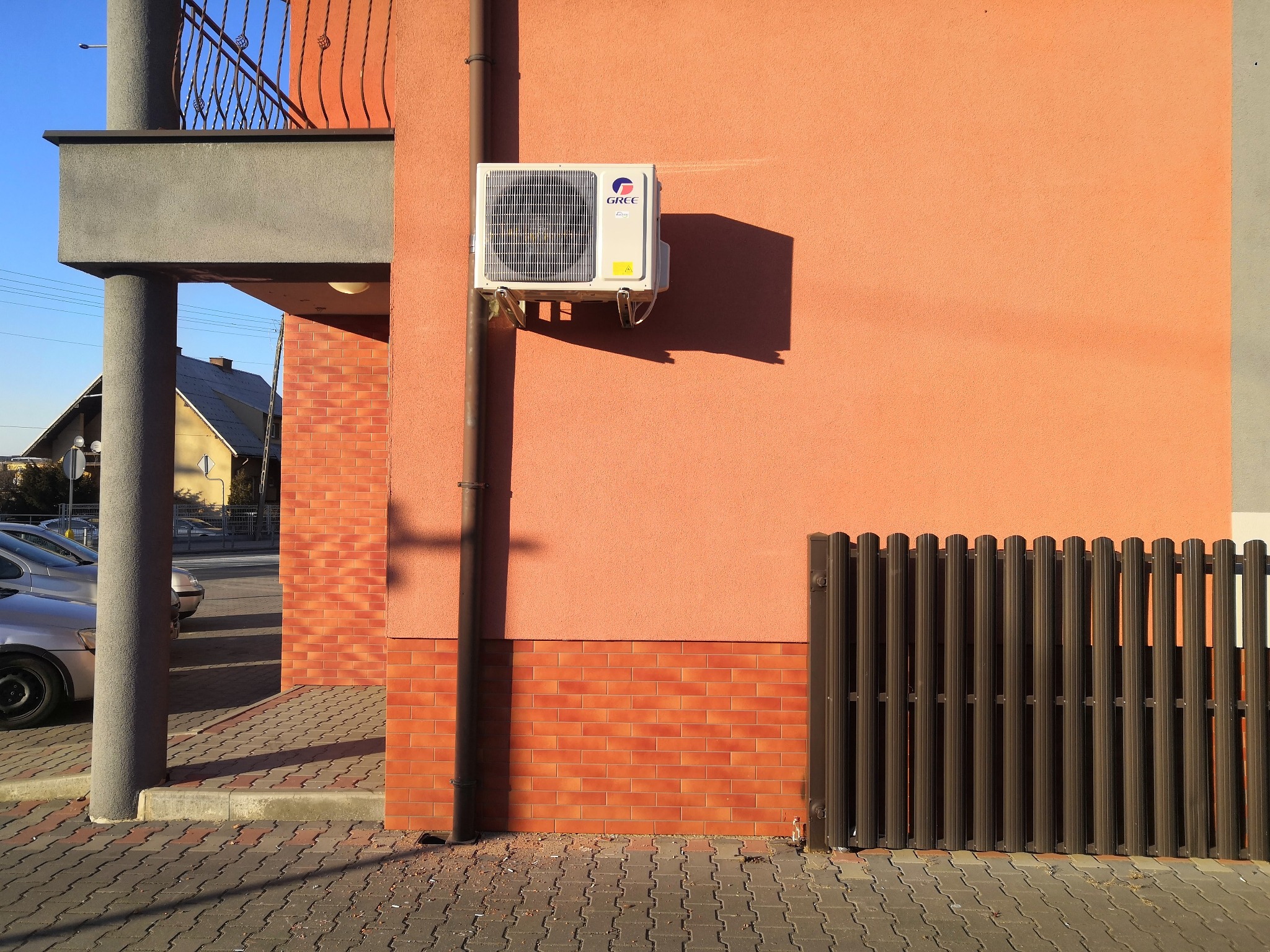 Biała jednostka klimatyzacyjna marki Gree zamontowana na pomarańczowej ścianie budynku, obok brązowej rynny spustowej i segmentu ogrodzenia.