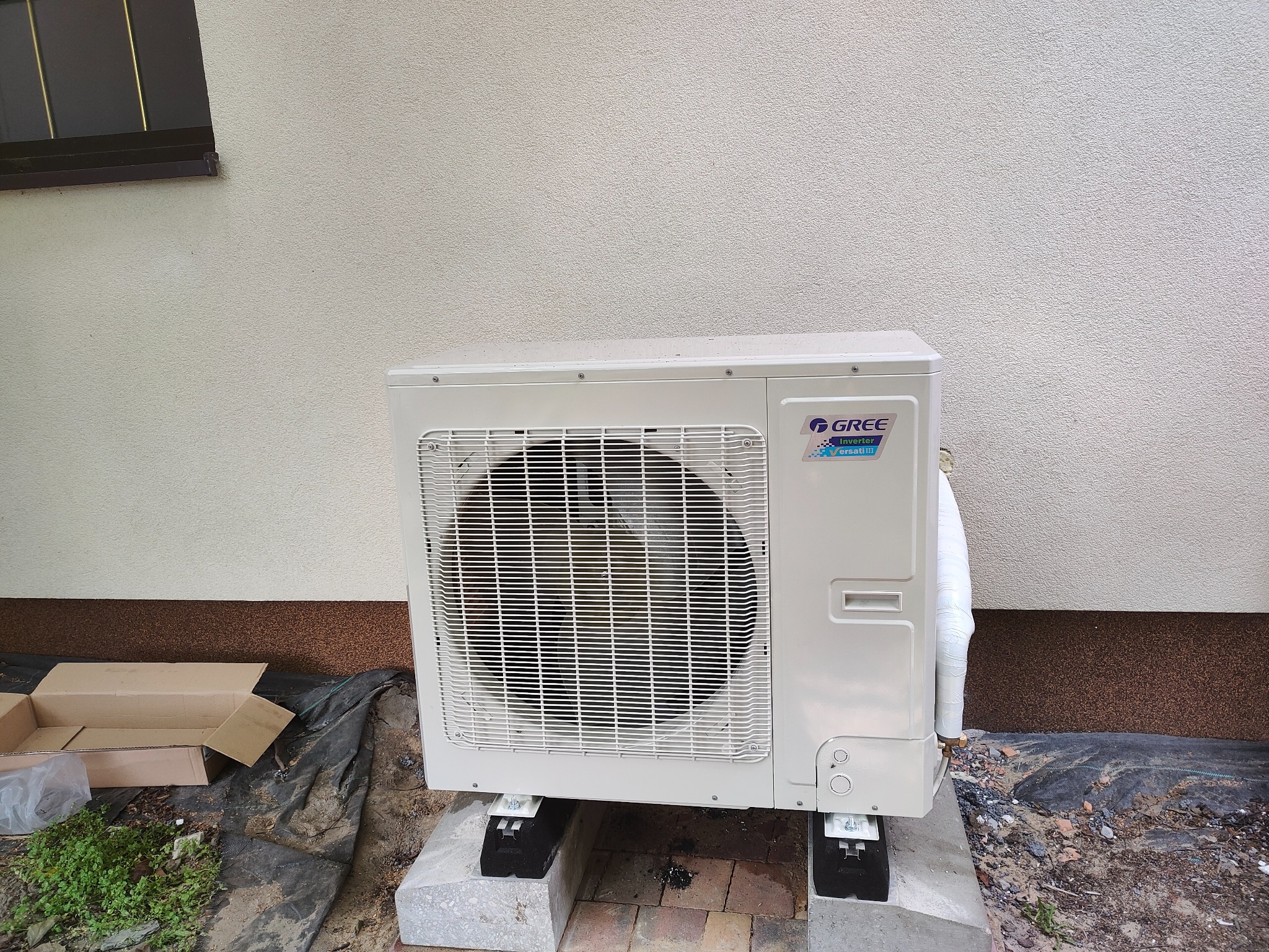 Zewnętrzna jednostka klimatyzacji marki Gree, zamontowana na betonowych podstawach z gumowymi podkładkami, obok ściany budynku z widocznym oknem i kartonem na ziemi.