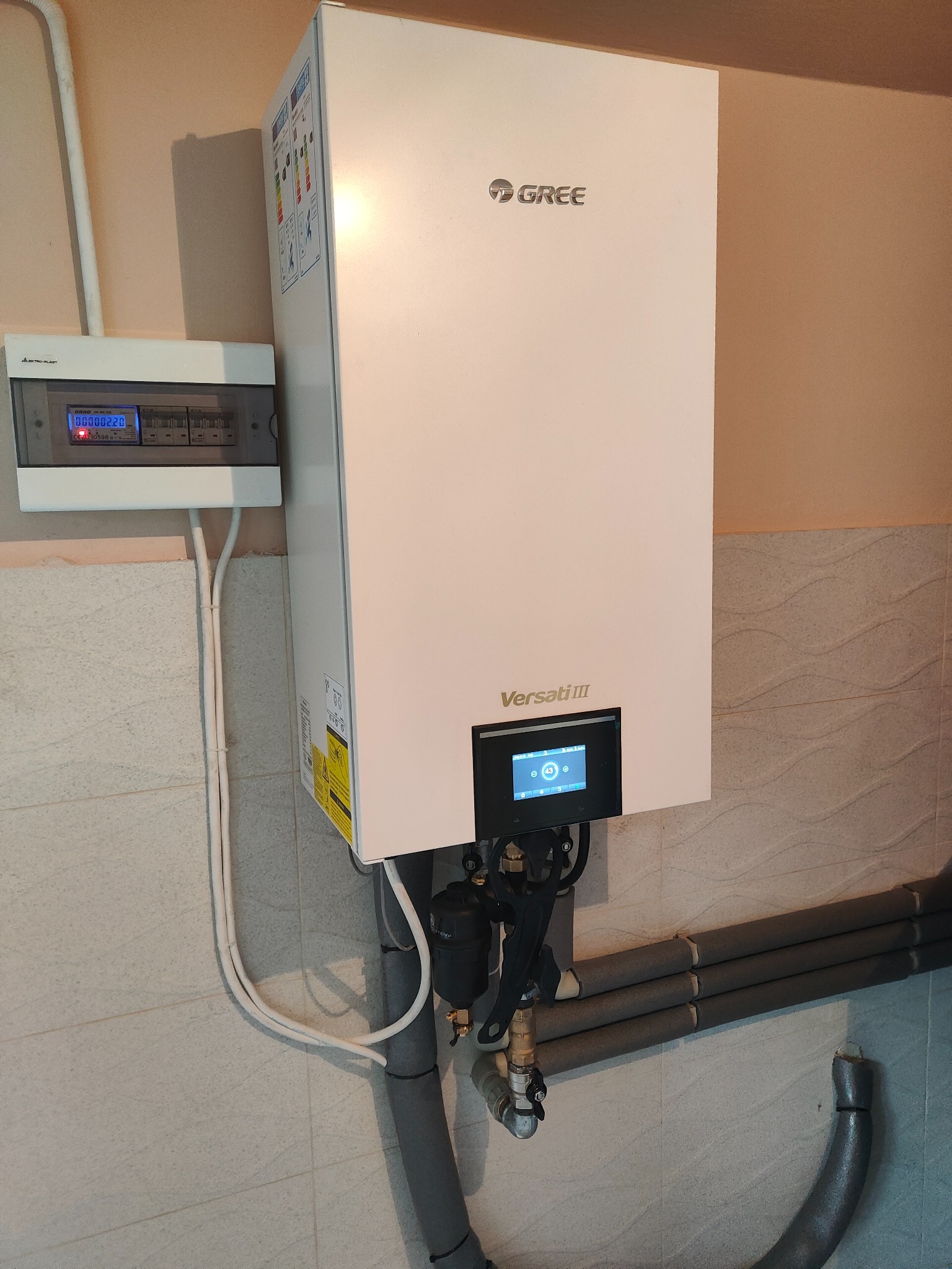 Biała pompa ciepła marki Gree Versati III zamontowana na ścianie z szarą płytką, obok skrzynka elektryczna z wyświetlaczem, widoczne rury z izolacją.