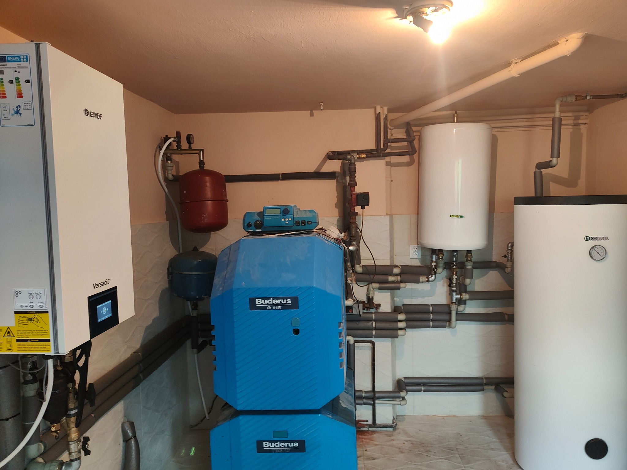 Instalacja centralnego ogrzewania z kotłem Buderus, zasobnikiem Kospel i pompą ciepła Gree Versati III w pomieszczeniu technicznym z rurami w izolacji.