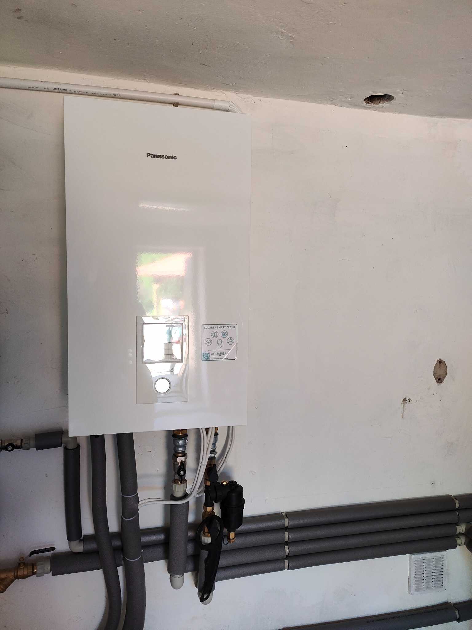 Biała jednostka Panasonic Aquarea Smart Cloud zamontowana na ścianie z widocznymi rurami w izolacji termicznej.