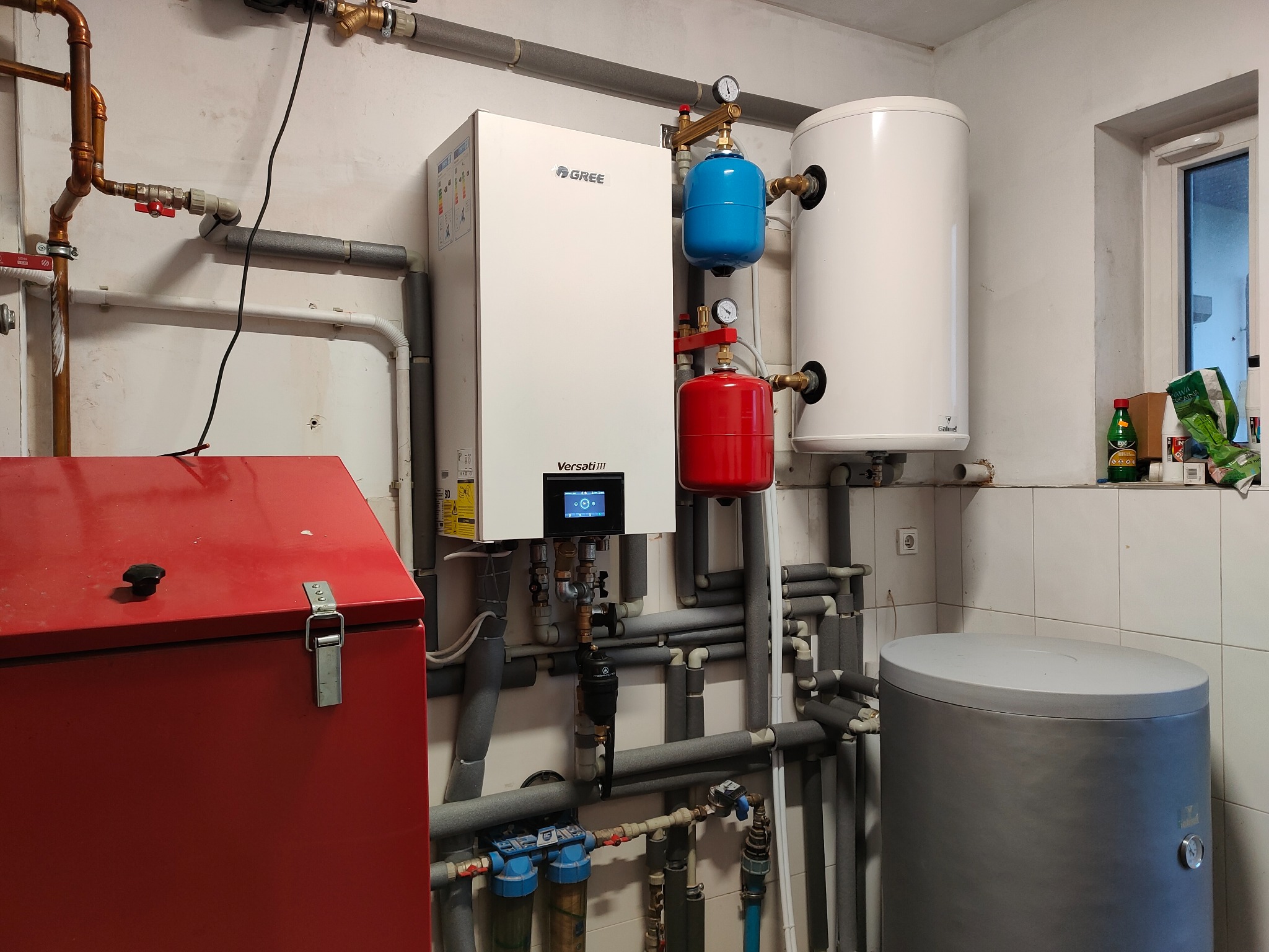 Pompa ciepła Gree Versati III z zbiornikiem buforowym, naczyniami przeponowymi w kolorach czerwonym i niebieskim oraz licznymi rurami, zainstalowana w pomieszczeniu z białymi płytkami i czerwonym...