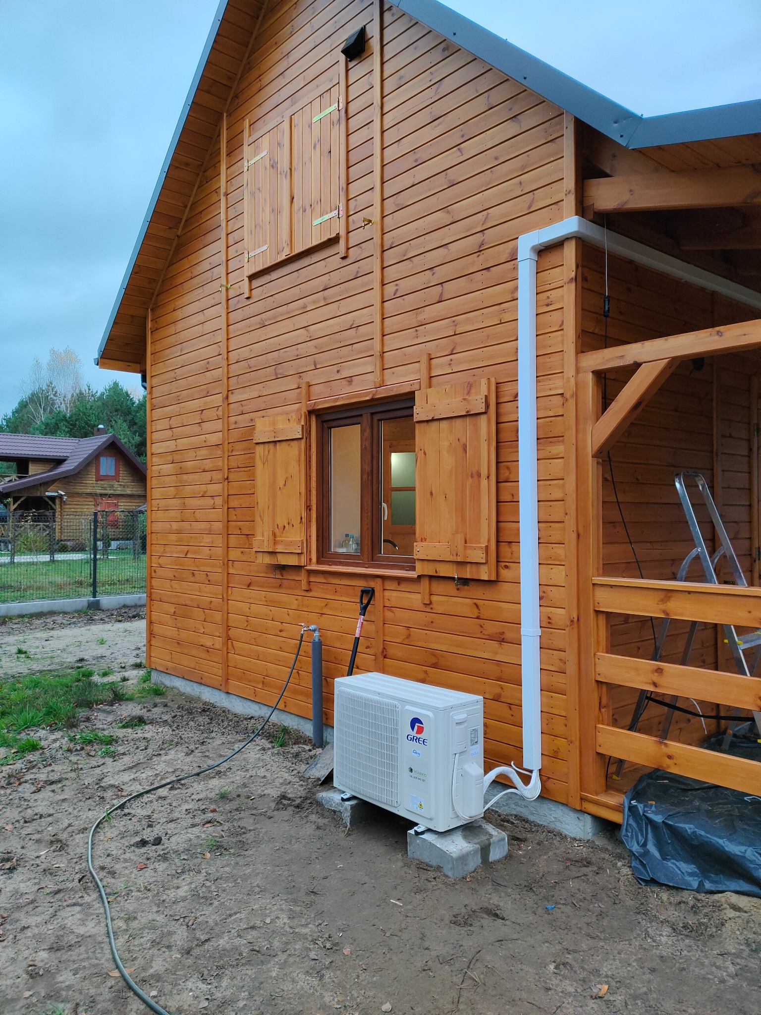 Biała jednostka zewnętrzna klimatyzacji marki Gree, umieszczona na betonowych bloczkach przy drewnianym domku z okiennicami, widoczny przewód odprowadzający skropliny i rura instalacyjna.