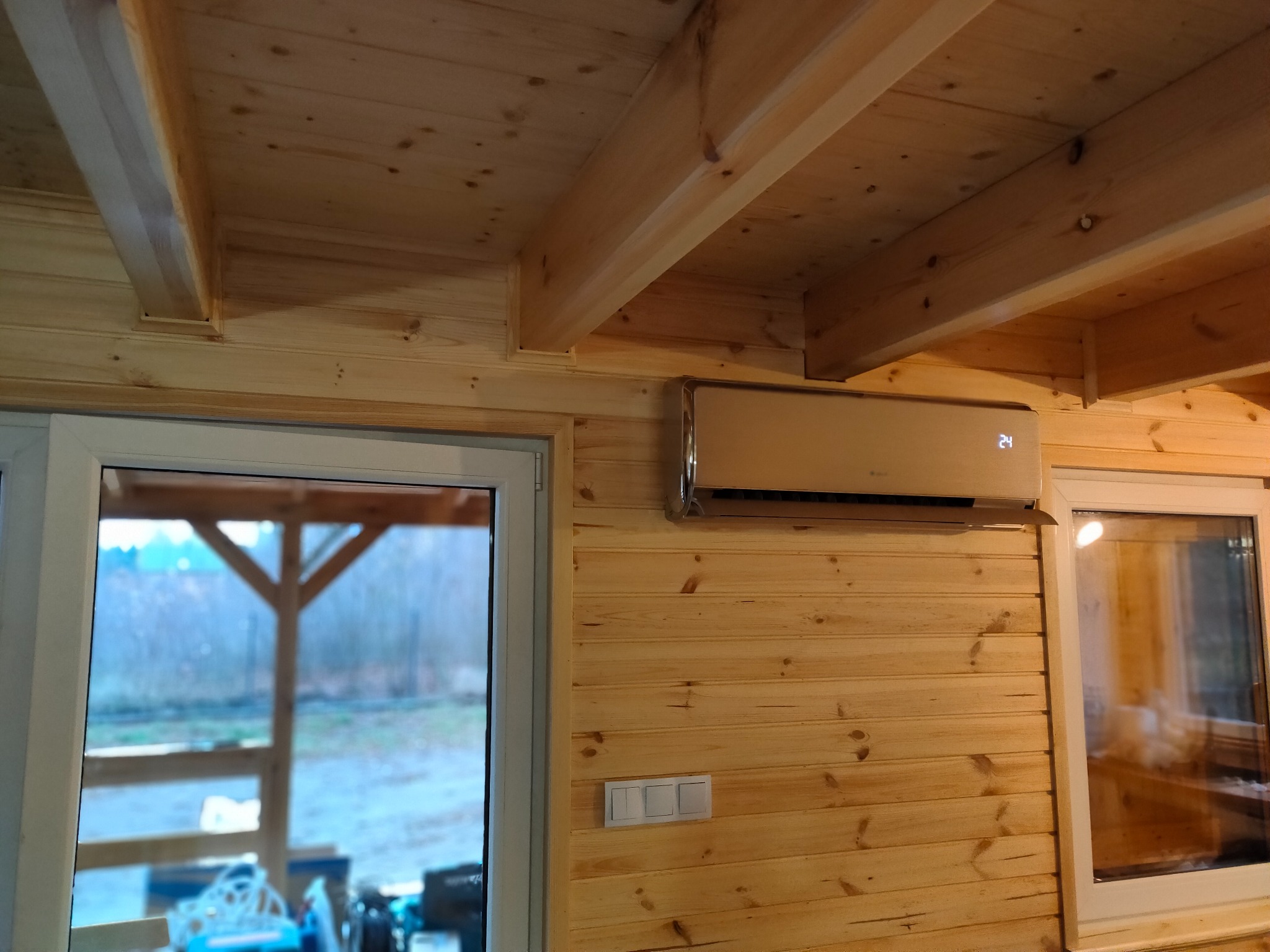 Klimatyzator ścienny w obudowie w kolorze szampańskim zamontowany w pomieszczeniu wykończonym drewnianą boazerią, widoczne okno z białymi ramami i drewniany sufit z belkami.