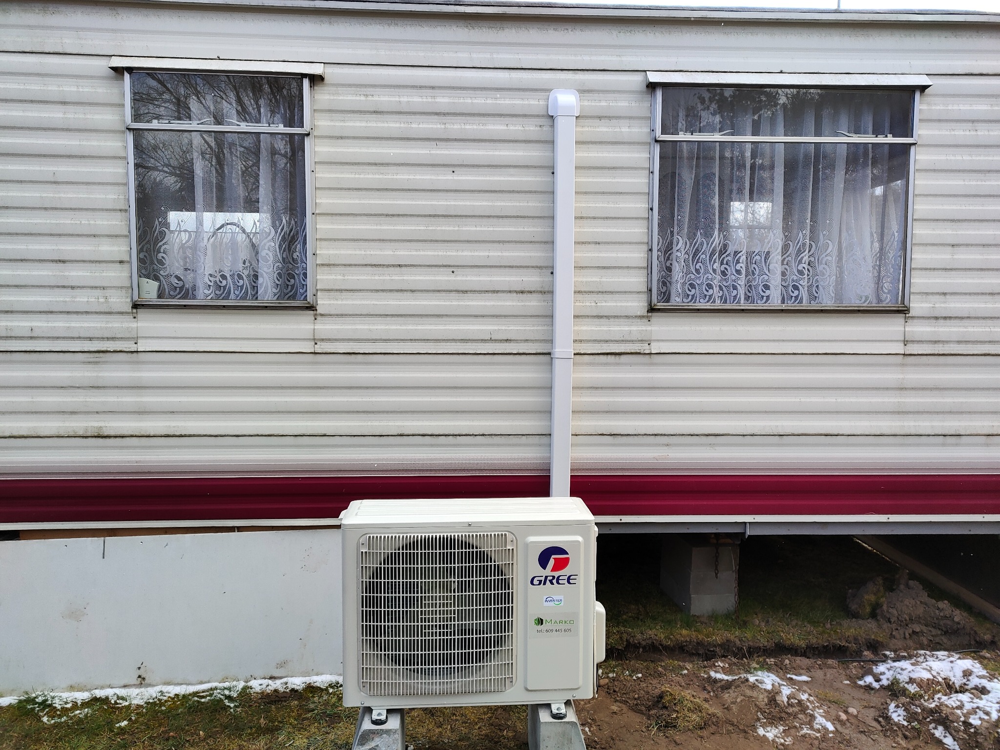 Biała jednostka zewnętrzna klimatyzacji marki Gree, zamontowana na betonowych podstawach przy ścianie budynku typu barakowóz z dwoma oknami z firankami.