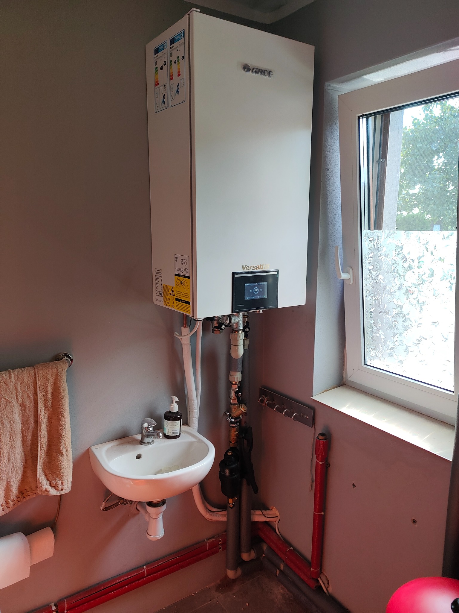 Biała pompa ciepła marki Gree zamontowana na szarej ścianie nad umywalką w małej łazience z oknem z ozdobną folią.