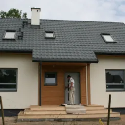 Nowo wybudowany dom z drewnianą elewacją, szarą dachówką i lukarnami, pracownik przy drzwiach wejściowych.