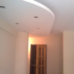 Wykończone wnętrze: biały sufit z podwieszaną, zaokrągloną konstrukcją i punktowym oświetleniem, ściany w jasnym odcieniu beżu, drewniane drzwi z szybkami prowadzące do innego pomieszczenia.