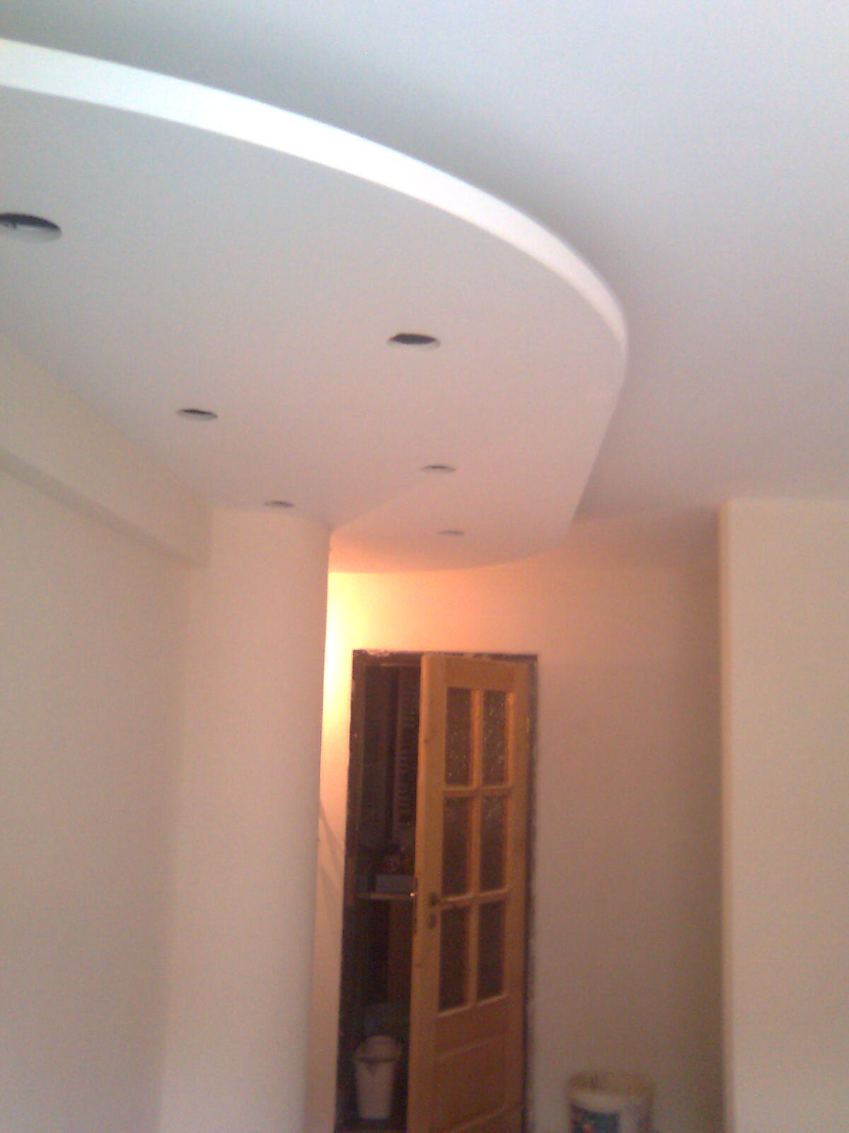 Wykończone wnętrze: biały sufit z podwieszaną, zaokrągloną konstrukcją i punktowym oświetleniem, ściany w jasnym odcieniu beżu, drewniane drzwi z szybkami prowadzące do innego pomieszczenia.