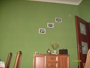 Ściana pomalowana na zielono z trzema zdjęciami w białych ramkach, komoda z dekoracjami, drewniane krzesła i drzwi. Widoczny fragment kanapy.