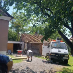 Pracownik w szarych spodenkach przycina drewno na zewnątrz, na tle remontowanego budynku z odkrytą konstrukcją dachu i zaparkowanym samochodem dostawczym z otwartymi drzwiami.