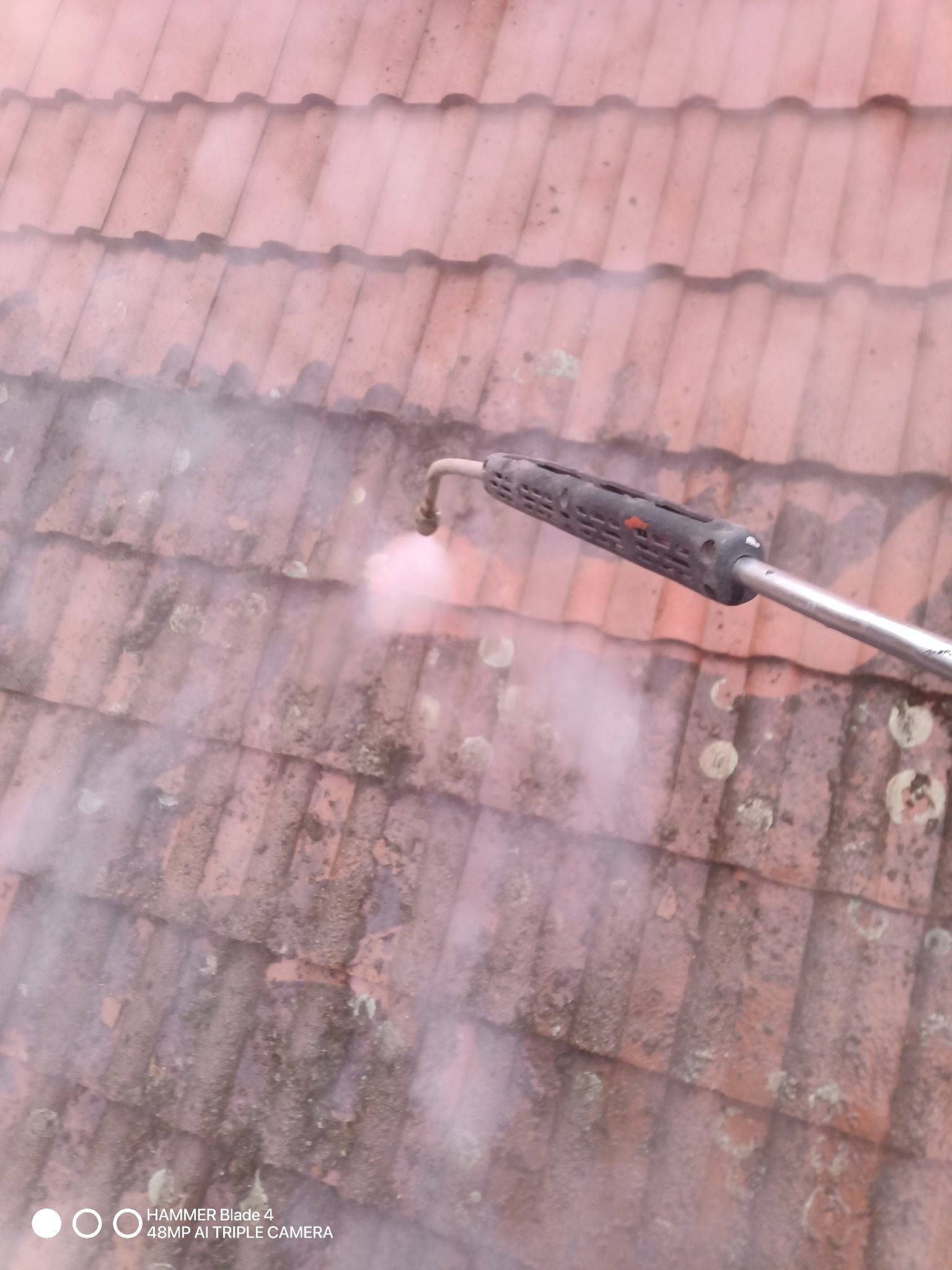 Czyszczenie dachówki ceramicznej za pomocą myjki ciśnieniowej, widoczne zabrudzenia i strumień wody pod ciśnieniem.