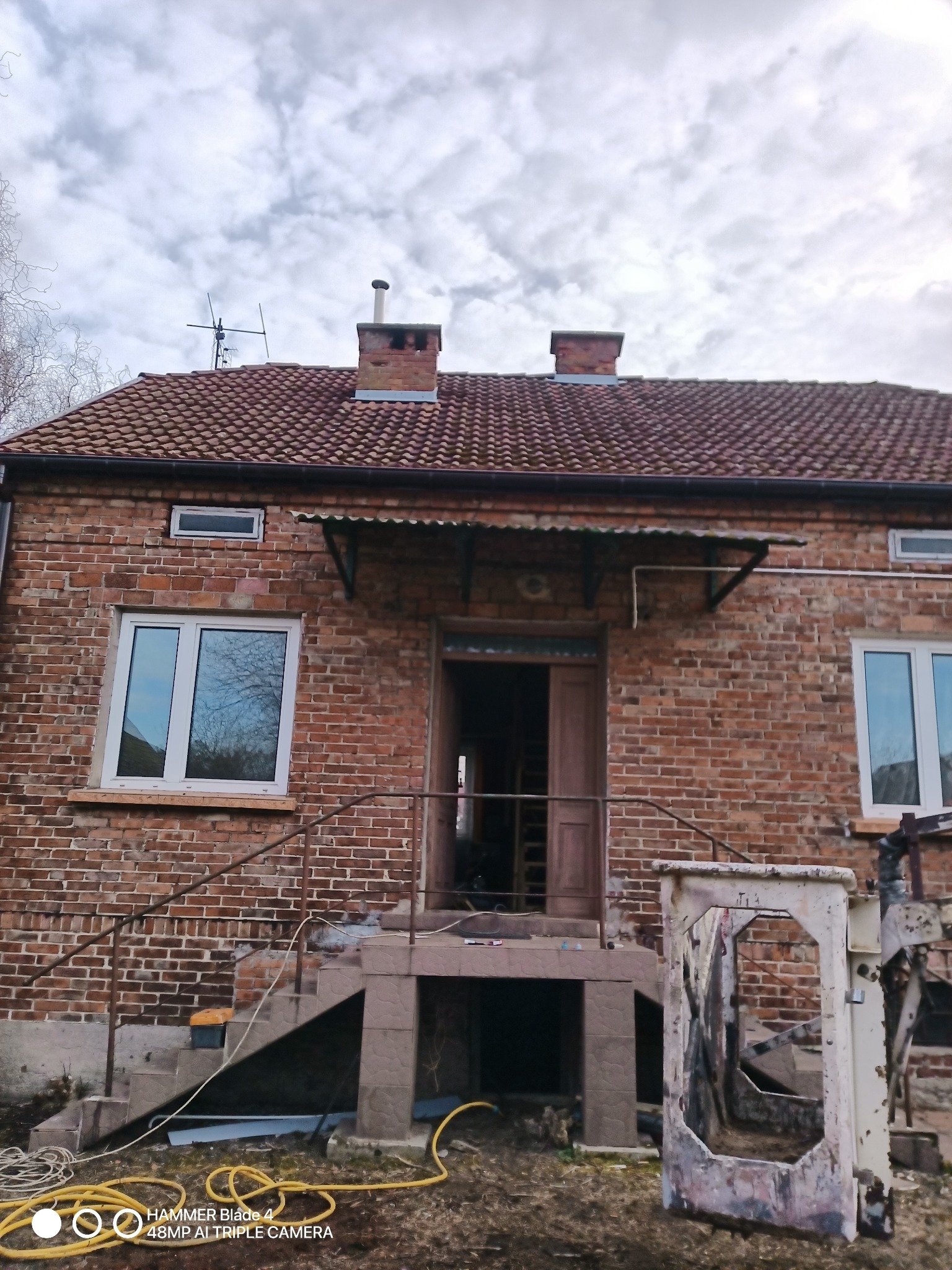 Ceglany dom z terakotową dachówką, widoczne schody wejściowe, okna z białymi ramami i częściowo odsłonięte poddasze z kominami, w tle pochmurne niebo.